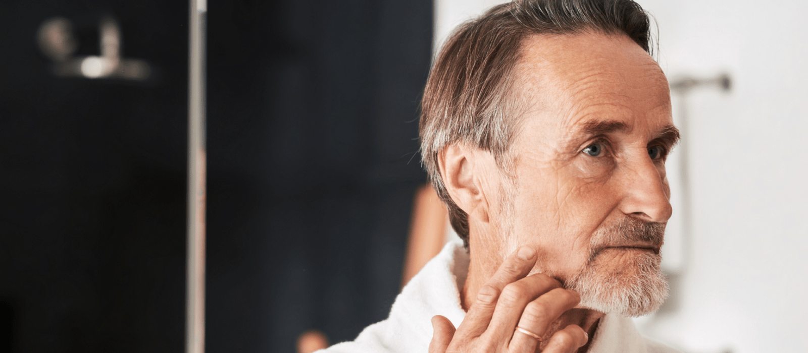 Senior man touching his face in bathroom in from of a mirror. Male in bathrobe looking at his reflection inspecting skin.